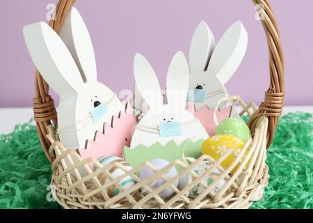 Coniglietti di legno con maschere protettive e uova dipinte in cestello su erba di carta, primo piano. Vacanze di Pasqua durante la quarantena COVID-19 Foto Stock