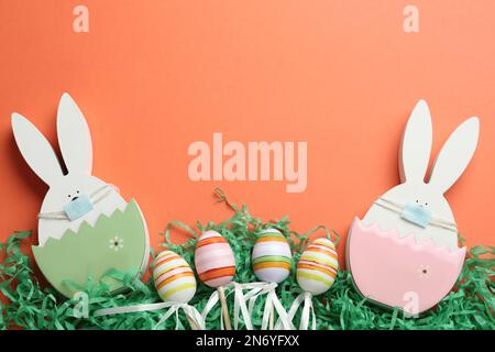 Coniglietti di legno con maschere protettive, uova e spazio per testo su sfondo corallo, giacitura piatta. Vacanze di Pasqua durante la quarantena COVID-19 Foto Stock