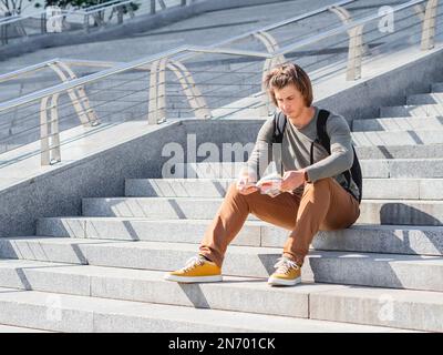 Il giovane si siede su una scala di pietra nel parco e legge la guida turistica. Viaggiare da solo in città. Turismo urbano. Monumenti architettonici moderni. Foto Stock