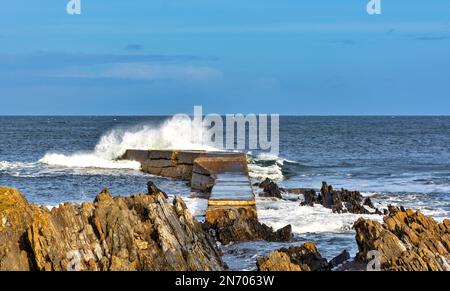 Il Sandend Village Aberdeenshire Scotland ondeggia e spruzza sopra il frangiflutti e le rocce in una giornata ventilata Foto Stock