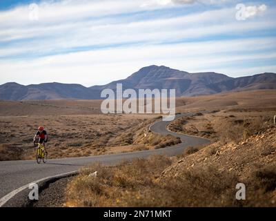 Ciclista stradale sulla strada secondaria tortuosa nella regione collinare centrale di Fuerteventura vicino a Juan Gopar, vista sulla montagna Montäa de Melindraga, Isole Canarie, Spagna Foto Stock
