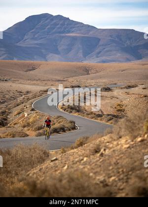 Ciclista stradale sulla strada secondaria tortuosa nella regione collinare centrale di Fuerteventura vicino a Juan Gopar, vista sulla montagna Montäa de Melindraga, Isole Canarie, Spagna Foto Stock
