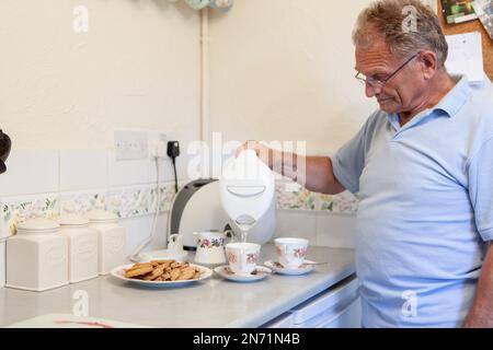 Pensione: Versare il tè. Un uomo anziano nella sua cucina che prepara uno standard britannico - 2 tazze di tè con biscotti. Da una serie di immagini correlate. Foto Stock