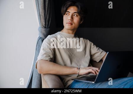 Giovane asiatico che lavora con il computer portatile mentre si siede in poltrona a casa Foto Stock