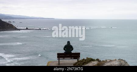 Uomo che guarda il mare in un posto bellissimo con le montagne su una panchina Foto Stock