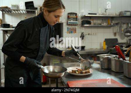 Giovane donna cuoca in cucina professionale mette la carne sul piatto Foto Stock