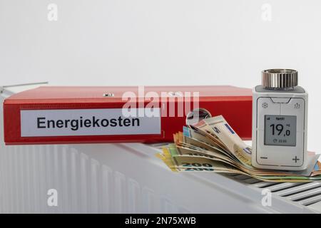 Cartella file rossa con etichetta costi energetici, termostato radiatore WLAN FRITZ! DECT 302, il display mostra 1ö°C., varie bollette in euro giacciono sul radiatore, 705 euro, immagine simbolo, costi energetici, costi di riscaldamento in aumento, networking, digitale, tecnologia per la casa intelligente, sfondo bianco, Foto Stock