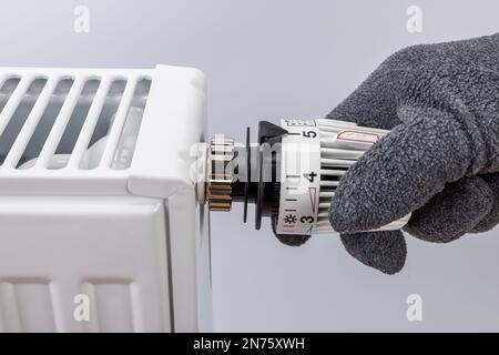 La mano dell'uomo con guanto aumenta la temperatura ambiente sul termostato del radiatore, impostando il livello 5 circa 2ö°C., sfondo bianco, immagine simbolo, costi energetici, costi di riscaldamento in aumento, Foto Stock