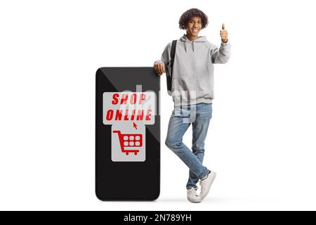 Giovane afro americano maschio gesturing pollici su e appoggiandosi su un grande telefono cellulare con negozio di testo online isolato su sfondo bianco Foto Stock