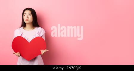 Concetto di San Valentino. Solitario adolescente asiatica ragazza sognando di amore, sentendosi triste e solitario in giornata degli amanti, guardando da parte con pietà, tenendo grande rosso Foto Stock