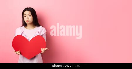 Concetto di San Valentino. Single teenage asian girl vuole innamorarsi, guardando triste e solitario alla macchina fotografica, sulking afflitto in festa degli amanti, tenendo grande Foto Stock