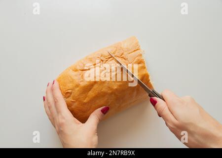 la mano femminile taglia un pezzo di pane con un coltello su un tavolo bianco in cucina a casa Foto Stock