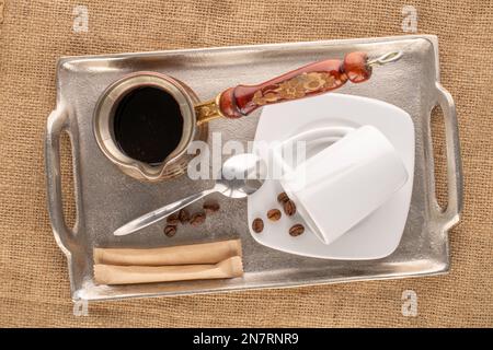 Una cezve con caffè, piattino bianco in ceramica con tazza, cucchiaio di metallo, diversi chicchi di caffè aromatici e due bustine di zucchero con vassoio di metallo su coagulo di iuta Foto Stock
