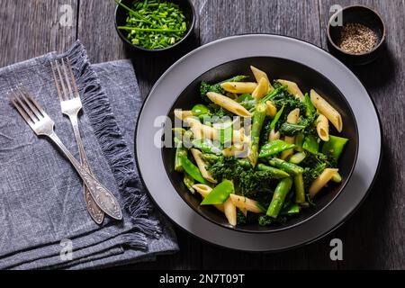 Pasta primavera con asparagi, piselli, broccoli e cavolo in ciotola nera su tavolo di legno scuro con forchette Foto Stock