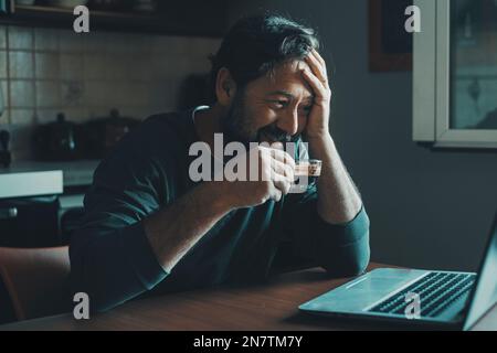 L'uomo da solo a casa usa un computer portatile sorridente e beve caffè espresso in cucina. Mattina presto lavoro e tecnologia internet indoor attività di svago c Foto Stock