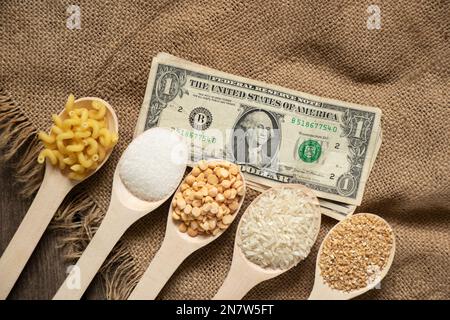 zucchero riso piselli pasta cereali su cucchiai di legno e dollari americani sul tavolo in cucina, cibo il prezzo del cibo Foto Stock