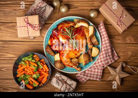 Cena natalizia con pollo intero arrosto e verdure vista dall'alto su sfondo rustico in legno con giocattoli natalizi e regali avvolti, festivo Foto Stock