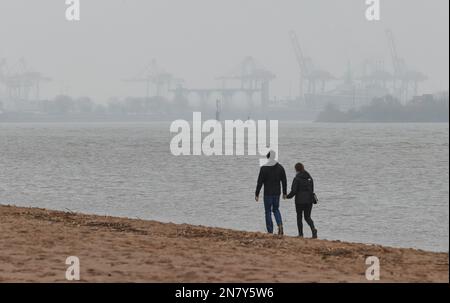 Amburgo, Germania. 11th Feb, 2023. Un uomo e una donna camminano tenendo le mani sotto il cielo coperto e piovono leggero sulla spiaggia di Elba a Övelgönne. Sullo sfondo si possono vedere gru e container dal porto di Amburgo. Credit: Georg Wendt/dpa/Alamy Live News Foto Stock