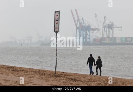 Amburgo, Germania. 11th Feb, 2023. Un uomo e una donna camminano tenendo le mani sotto il cielo coperto e piovono leggero sulla spiaggia di Elba a Övelgönne. Sullo sfondo si possono vedere gru e container dal porto di Amburgo. Credit: Georg Wendt/dpa/Alamy Live News Foto Stock