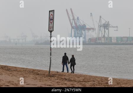 Amburgo, Germania. 11th Feb, 2023. Un uomo e una donna camminano tenendo le mani sotto il cielo coperto e piovono leggero sulla spiaggia di Elba a Övelgönne. Sullo sfondo si possono vedere gru e container dal porto di Amburgo. Credit: Georg Wendt/dpa/Alamy Live News Foto Stock