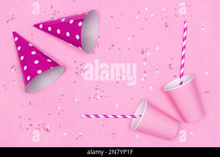 Tazze di carta, cannucce da bere a righe, cappelli da festa con spruzzate di caramelle su sfondo rosa. Concetto di festa di compleanno. Spazio di copia Foto Stock