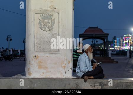 Diu, India - Dicembre 2018: Un uomo musulmano indiano anziano seduto accanto ad un'antica colonna bianca con incisioni e testi dell'era portoghese la sera. Foto Stock