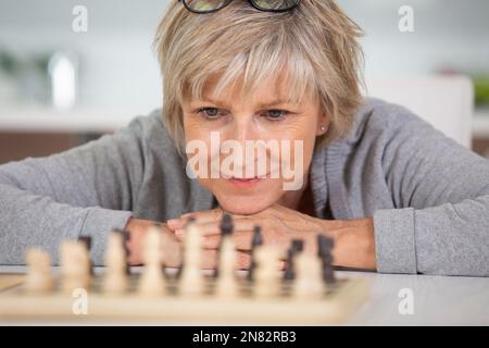 donna anziana e penitente seduta al tavolo e giocando a scacchi Foto Stock