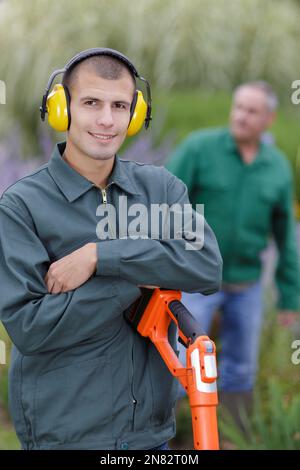 Giardiniere sorridente in uniforme, occhiali protettivi, cuffie e guanti  che tengono tagliasiepi a benzina mentre si trova sul retro del cortile.  Uomo caucasico che utilizza attrezzature da giardinaggio moderne all'aperto  Foto stock 