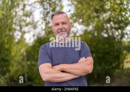 Uomo sicuro di mezza età in piedi davanti agli alberi verdi con le braccia incrociate Foto Stock