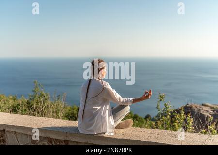 Profilo di una donna che fa yoga in cima a una scogliera in montagna. Donna medita nello yoga asana Padmasana. Foto Stock