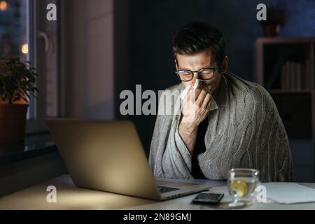 Uomo d'affari malato che soffia il naso mentre lavora al computer in ufficio a casa di notte. Il giovane uomo starnutisce con il naso che corre mentre lavora da casa in auto-isolamento, ha freddo cattivo, influenza, febbre. Rimani a casa Foto Stock