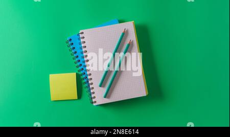 Beffa. Un taccuino A4 vuoto con una molla a spirale, matite e adesivi su sfondo verde, vista dall'alto, piatto Foto Stock