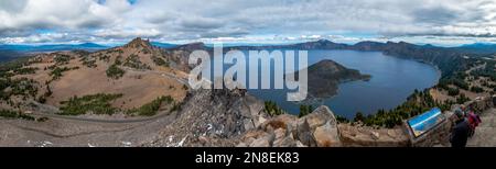 Vista panoramica del Crater Lake con Wizard Island al centro e un cielo con molte nuvole. La strada che la circonda e il punto panoramico con un po' di tour Foto Stock