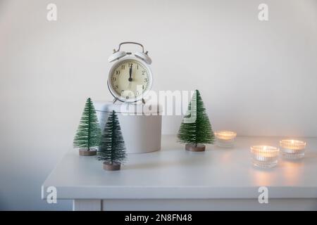 Una sveglia bianca, un puntatore a dodici, un piccolo albero di Natale artificiale verde e candele accese in candelabri su una cassettiera bianca. Copia Foto Stock