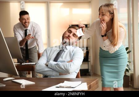 Giovane donna che si attacca nota con parola Fool al volto del collega in ufficio. Scherzo divertente Foto Stock