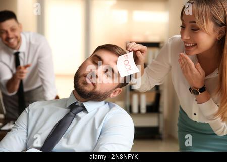 Giovane donna che si attacca nota con parola Fool al volto del collega in ufficio. Scherzo divertente Foto Stock