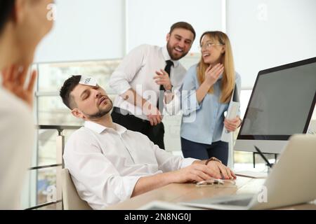 I colleghi prendono nota con la parola Fool al volto del collega in ufficio. Scherzo divertente Foto Stock