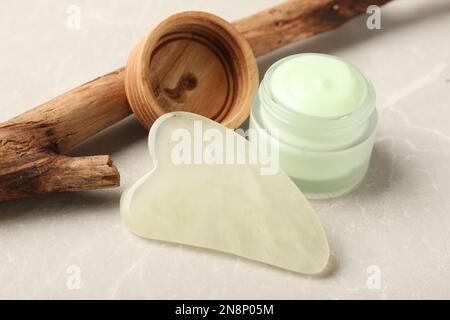 Jade gua sha strumento, vaso di crema e ramo d'albero su tavolo grigio Foto Stock