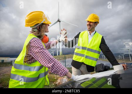 Due colleghi soddisfatti si danno a vicenda un alto cinque mentre lavorano consultando un disegno tecnico in una fattoria solare. Foto Stock