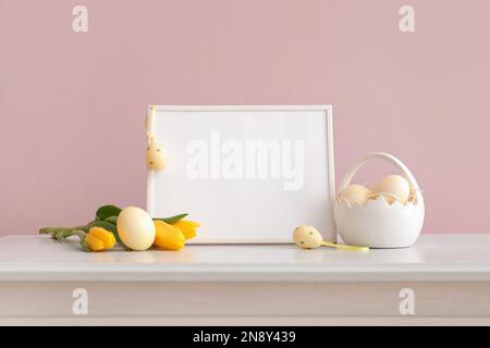 Cornice vuota, cestino con uova di pasqua dipinte e fiori di tulipano giallo sul cassetto vicino alla parete rosa Foto Stock