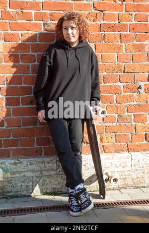 Una ragazza adolescente con capelli ricci vestiti di nero si alza con un longboard vicino ad un muro di mattoni Foto Stock