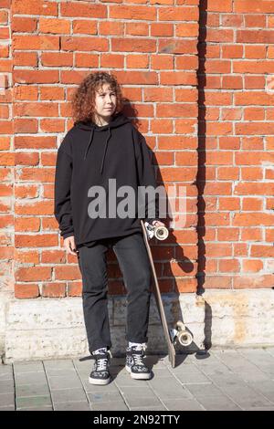 Una ragazza adolescente con capelli ricci vestiti di nero si alza con un longboard vicino ad un muro di mattoni Foto Stock