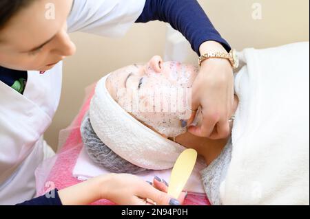 Un estetista rimuove una maschera idratante del foglio sul viso di una donna con una spatola, procedure cosmetologiche. Foto Stock