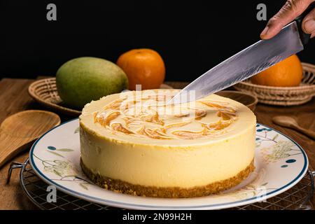 Uomo che usa un coltello che taglia la cheesecake fatta in casa di mango in un piatto di ceramica su una griglia di metallo con mango, arance mature, cucchiaio di legno, tazza e vassoio di bambù Foto Stock