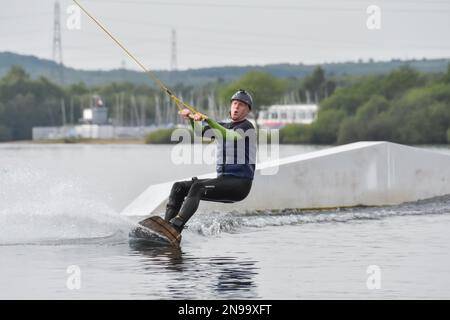 Staffordshire, Regno Unito. Maggio 2018. Wakeboarder surf attraverso il lago utilizzando un cavo di traino Foto Stock