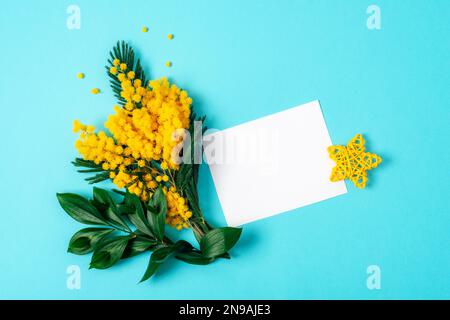 Scheda vuota e mimosa fiori gialli su sfondo blu. Concetto di vacanze primaverili. Vista dall'alto, piatto, mockup. Foto Stock