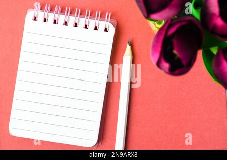 Quaderno a spirale aperto con carta rigata e matita con fiore di tulipano rosso su sfondo rosso. Foto Stock
