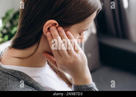 Primo piano di una giovane bruna femmina che tiene l'orecchio doloroso, improvvisamente sensazione di forte dolore. Donna caucasica malsana 20s che soffre di otite dolorosa sitti Foto Stock