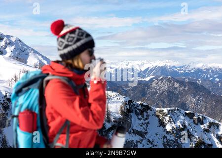 Un'escursionista turista femminile beve tè caldo durante un'escursione invernale sul monte Dobratsch, godendosi spettacolari viste panoramiche innevate in Austria. Foto Stock