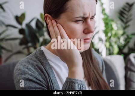 Primo piano del profilo laterale di una giovane femmina caucasica malata con dolore all'orecchio che tocca la testa dolorosa seduta sul divano. Donna malsana 20s che soffre di dolore Foto Stock